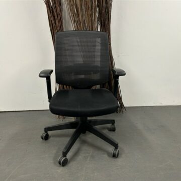 A Black Operator Chair with Grey Flecked Mesh Back featuring armrests and five caster wheels sits on a gray floor before a white wall, with tall decorative brown branches placed behind it.