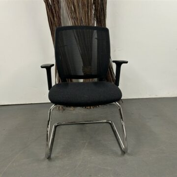 A Meeting Chair with Chrome Cantilever Frame, featuring a black mesh back and cushioned seat, is placed on a gray floor against a white wall, with tall decorative sticks in the background.
