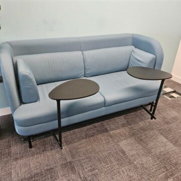 A light blue sofa with high arms sits against a light blue wall, with two black, leaf-shaped side tables positioned in front of each seat. The floor is covered with gray carpet tiles.