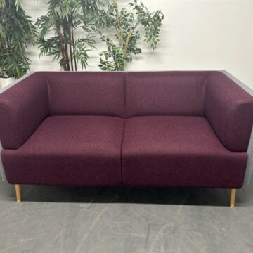 A modern two-seater sofa with maroon upholstery and gray armrests, set on wooden legs. Green potted plants are in the background against a plain white wall.