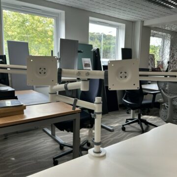 Dual monitor arm mounts attached to a desk in a modern office with several empty workstations, office chairs, and large windows letting in natural light.