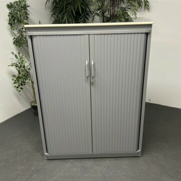A gray metal cabinet with two vertical handles and ribbed doors stands on a dark floor, with potted green plants in the background against a white wall.