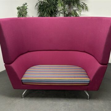 A modern, magenta high-back sofa with a striped cushion sits on a gray floor with green plants in the background against a plain white wall.