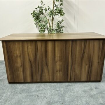 A rectangular wooden desk with a natural wood grain finish stands on a gray carpeted floor. A leafy green plant is positioned behind the desk against a plain white wall.