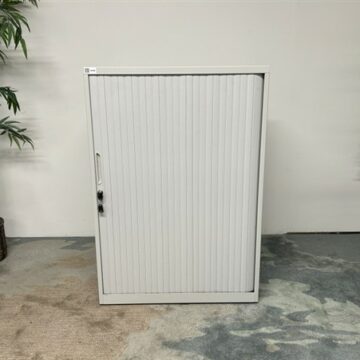 A white metal storage cabinet with a vertical tambour door stands against a white wall, flanked by two potted plants on either side, on a carpeted floor with varying shades.