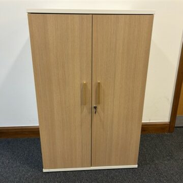 A wooden cabinet with two vertical doors and silver handles, standing against a white wall on a dark carpeted floor. One of the handles has a key inserted into its lock.