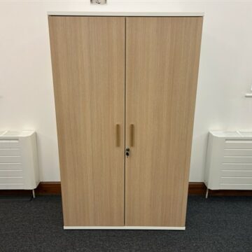 A wooden cabinet with two closed doors and metal handles stands against a white wall between two windows, above a dark carpeted floor.