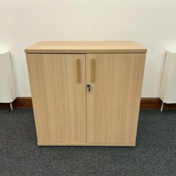 A light wood cabinet with two doors and a lock in the center, standing on a dark carpeted floor against a white wall.
