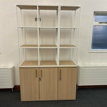 A wooden cabinet with three doors at the bottom and an open white metal shelving unit with nine compartments on top, placed against a white wall between two windows.