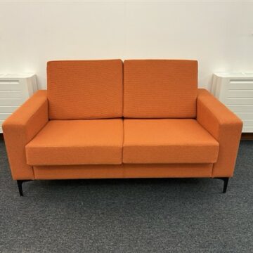 A bright orange two-seater sofa with straight arms and black legs, placed on a gray carpet in a room with white walls and radiators beneath two windows.