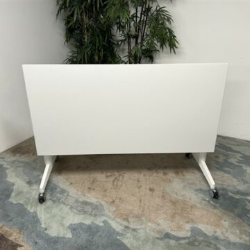 A white rectangular table on wheels is positioned in front of a green potted plant, set against a plain white wall and a carpet with blue, beige, and brown patterns.
