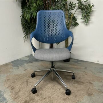A modern office chair with a blue perforated plastic backrest, grey cushioned seat, and silver metal swivel base with black wheels, positioned on a patterned carpet near green plants.
