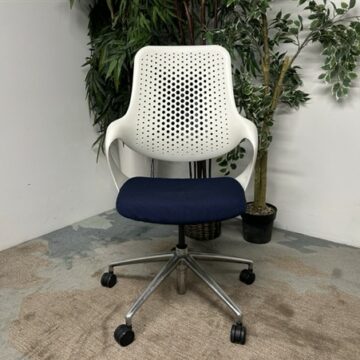 A modern white office chair with a perforated backrest, blue cushioned seat, and metal base on wheels is placed on a carpeted floor near green potted plants against a white wall.