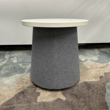A small round table with a white top and a gray, fabric-covered cylindrical base sits on a patterned carpet in front of a plain wall.
