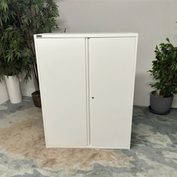 A white metal storage cabinet with two doors stands on a carpeted floor, flanked by two green potted plants against a pale wall.