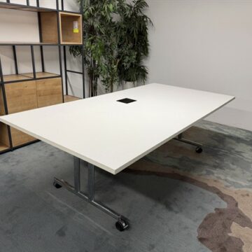 A large, white rectangular conference table on wheels stands in an office with a gray carpet. Wooden shelves and two potted plants are visible in the background.